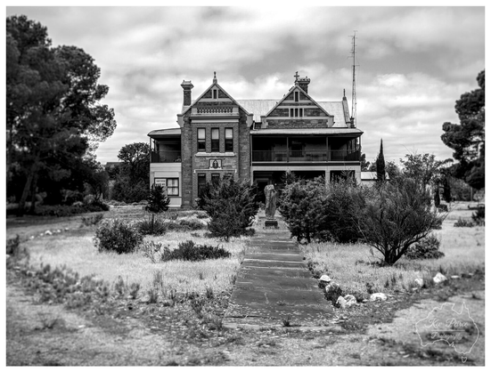 A black and white photo of the historic and imposing two story St. Cecilia's building in Peterborough, South Australia.

The grand brick structure features Victorian architectural details, including a wrap around veranda on the lower level and two prominent gables on the upper level.

A statue of a saint stands on the overgrown lawn, marking the beginning of a cracked, worn path leading directly to the front entrance.

The property is framed by mature trees on the left and right, set against a dramatic, cloudy sky.