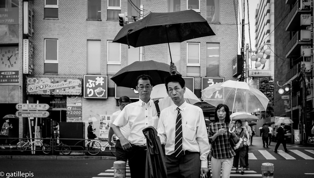 Dos salaryman travessant el carrer, un protegeix l'altre de la pluja amb un paraigua.