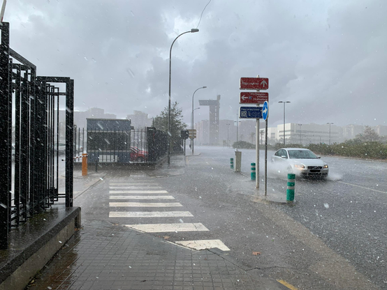 Fuerte tormenta en una calle urbana. Un coche se desliza por el agua, un paso de peatones mojado en primer plano, cielo nublado y señales de tráfico visibles. Al fondo la "Torre Miramar" de València (que parece una torre de control de una cárcel).