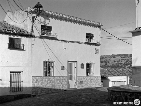 A monochrome image of a quaint house with a stone foundation and white plastered walls. The building features windows with bars and a wooden door, set against a backdrop of rolling hills and electric lines. A rustic lantern is fixed to the house.