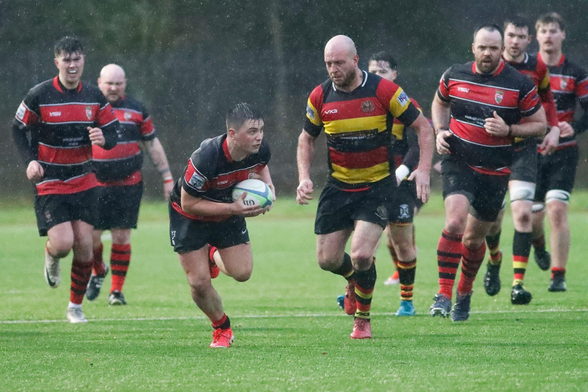 A rugby match is taking place in rainy conditions. Players in red and black uniforms are actively engaged, with one player running with the ball while others are in pursuit. The scene captures the intensity and action of the game.
