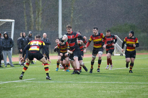 A rugby match in the rain, featuring players in red, black, and yellow jerseys competing for the ball. A player is sprinting while holding the ball, surrounded by teammates and opponents. Spectators are visible in the background, sheltered under umbrellas.
