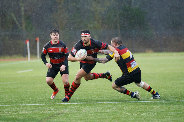 A rugby match in rainy conditions, featuring a player in a black and red uniform running with the ball, while two opponents attempt to tackle him. The scene captures the intensity of the game on a green field.
