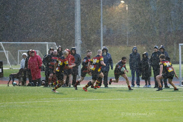 A rugby match is taking place in heavy rain, with players in colorful uniforms running on the field. Spectators in raincoats are watching from the sidelines. The scene captures the intensity of the game amidst challenging weather conditions.