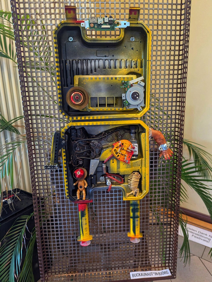 This image features a creatively repurposed toolbox transformed into a robot sculpture, mounted on a metal grid background. The top section of the toolbox serves as the robot’s head, complete with two distinct eyes. The bottom section forms the robot’s body, equipped with arms and legs constructed from an assortment of mechanical parts, including springs, gears, dolls, syringes, a glass bottle, and wires.

The sculpture is titled "Buckaunaut*in: Björg", as indicated by a sign on the bottom right. In the background, there is a blurry sign in German that reads "Thank you for supporting this festival!". The setting appears to be indoors, with a potted plant visible to the left.