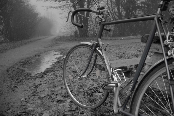 Das Schwarz-Weiß-Foto zeigt ein Fahrrad am Rande eines nebligen Weges. Der Weg führt in eine verschwommene, düstere Landschaft. Das Fahrrad lehnt an einer Holzbank und ist nur teilweise im Bild. Die Fotografie erzeugt eine melancholische und ruhige Stimmung.