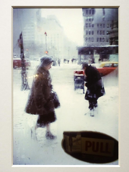 Een kleurenfoto uit New York na de tweede wereldoorlog van een straat waar het hartje winter is. De lens is ietwat beslagen door de vrieskou doordat de fotograaf achter een winkelruit deze opname heeft gemaakt. Een oudere vrouw passeert de winkelruit en een man probeert zijn handen warm te krijgen. In de verte zie je hoge gebouwen. Overal ligt sneeuw en de koud is voelbaar. De kleuren zijn daardoor ook gedempt. 