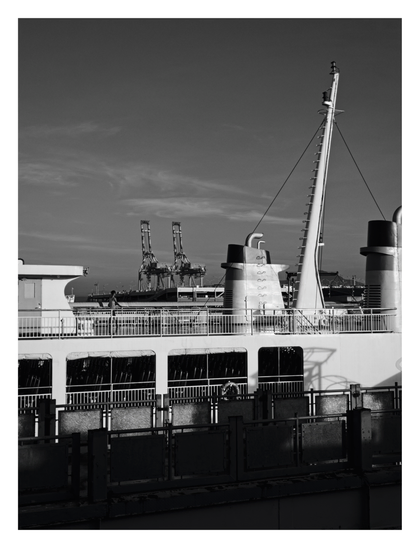The upper deck of a large white ship, featuring railings, two distinctive smokestacks, and a tall, angled mast. A lone figure stands at the ship's railing on the left side. In the distance, two large industrial gantry cranes rise against a sky textured with wispy clouds. The foreground is framed by a dark, silhouetted fence or barrier along the dock. - Google Gemini 3 Pro Preview
