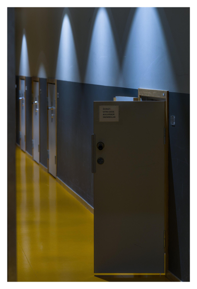 En stående bild på en korridor med dörrar in till förråd. Golvet är brunt, väggen mörkblå på nederdelen och ljusblå på överdelen. Den närmaste dörren står öppen. Lampor på väggen lyser i ett mönster på väggen.