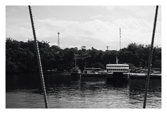In the center, a large ferry named "Santa Margarita" is docked at a pier alongside a smaller, darker vessel. The background consists of a dense, tree-covered hill rising from the shore. At the top of the hill, silhouetted against a cloudy sky, are communication towers and a prominent statue of a cross overlooking the bay. The water in the foreground is calm. - Google Gemini 3 Pro Preview