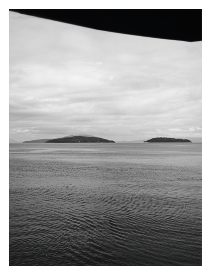 The foreground is filled with dark, rippling water that gradually lightens as it stretches toward the horizon. In the distance, two dark, tree-covered islands sit on the horizon line—a longer landmass on the left and a smaller, more distinct island on the right. Faint outlines of mountains are visible in the far background. Above, the sky is overcast with layers of soft, gray clouds. A dark, blurry shape intrudes at the very top of the frame, likely an overhang or part of a structure from where the photo was taken. - Google Gemini 3 Pro Preview