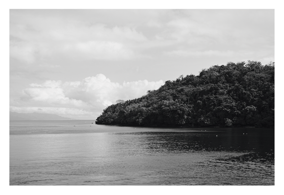 A calm body of water stretching out toward a distant horizon, flanked on the right by a large, densely forested hill or headland. The sky is filled with expansive, billowy clouds, and faint outlines of mountains are visible in the far distance. - Google Gemini 3 Pro Preview