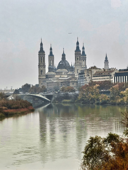 Fotografía en color. Se observa la Basílica del Pilar de Zaragoza al fondo, parcialmente envuelta en una neblina suave. En primer plano, el río Ebro refleja sutilmente los tonos grisáceos del cielo y los colores otoñales de los árboles. La imagen transmite una atmósfera serena y ligeramente melancólica.

Color photograph. The Basilica of Our Lady of the Pillar in Zaragoza stands in the background, partially veiled by soft mist. In the foreground, the Ebro River subtly reflects the gray sky and the autumn colors of the trees. The scene conveys a calm and slightly melancholic atmosphere.