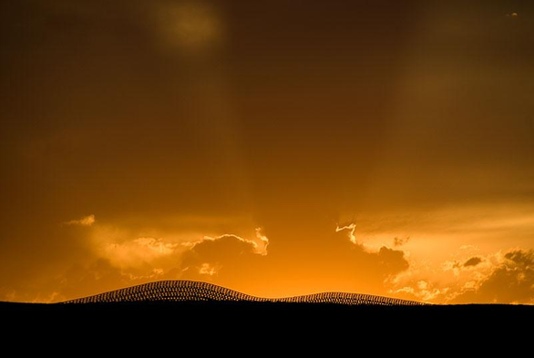 A very dramatic orange and yellow sunset over what looks like undulating hills, but on closer inspection the curves are snow fences rising over a flat ridge line. Behind them are scattered storm clouds and overhead is a thin, high layer of clouds. The Sun hitting towering cumulus casts a shadow on the clouds overhead which looks like a dark ray across the sky. 