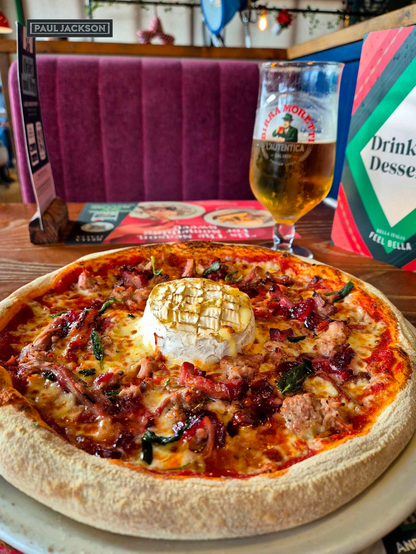 Christmas Camembert Pizza.

A delicious and very festive lunch, focusing on a close-up, top-down shot of a seasonal pizza. The pizza itself is beautifully prepared, featuring a well-baked crust and a generous amount of rich, seasonal toppings. These toppings include tomato & mozzarella cheese base, pulled turkey, pancetta and cranberry sauce. The star of the show is a distinctive, round of baked Camembert cheese sitting prominently right in the center,  with a golden-brown top which elevates the pizza to a decadent level.

In one corner of the image, there's the photographers name that reads "PAUL JACKSON" in white text on a dark rectangular background.

The scene is set on a wooden table inside a cozy restaurant. To complete the meal, a tall, stemmed glass of Birra Moretti beer is visible behind the pizza. The background includes a dark purple upholstered booth seat and a portion of a menu, collectively creating a warm, inviting, and slightly festive atmosphere for the dining experience.

Let the festive lunchtime indulgence commence!
