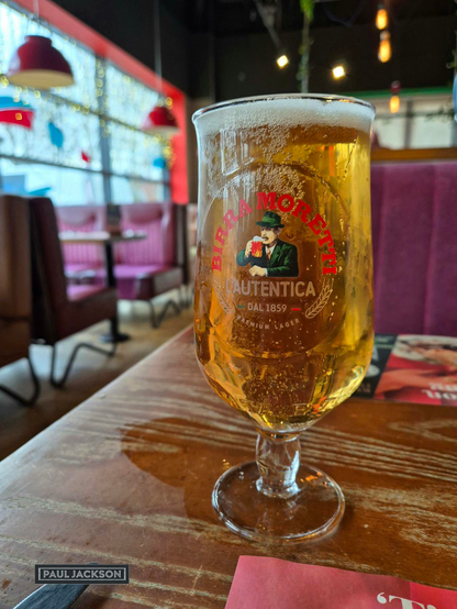 A close-up shot that captures a stemmed glass of Birra Moretti. The glass is filled with a clear, golden lager topped with a frothy white head, showing many small bubbles clinging to the sides. The center of the glass prominently features the distinctive Birra Moretti L'Autentica logo, complete with the image of the man in the green hat. The glass is resting on a textured wooden table. In the softly blurred background, a window provides daylight, and the bright lighting of the restaurant interior is visible, including red-shaded hanging lights, deep purple upholstered booth seating, and Christmas lights which add a rich, colorful warmth to the overall atmosphere.

In one corner of the image, there's the photographers name that reads "PAUL JACKSON" in white text on a dark rectangular background.

