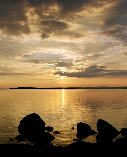 A sunset view over a broad lake. Calm waters reflect the setting sun. There are some rocks in the front. The sky is partially overcast.