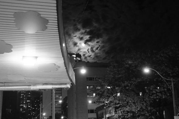 Just outside Wellesley Station, looking up at the night sky. The moon illuminates the soft curved shapes of the clouds. We see the overhang of the station, brightly lit, along the upper left. The right side of the frame is filled with the light and shadows of the street.
