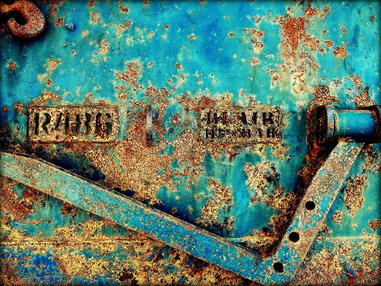 A colour photograph of a trawler rusted steel plate with R486 and Blair Dunbar text central on a background of weathered and rusted turquoise. An L-shaped arm lies diagonally across the bottom of the frame.