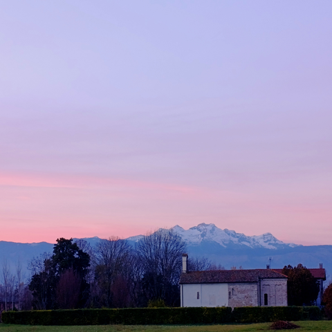 Tramonto sopra una piccola chiesa di campagna, attorniata da alberi. Il cielo è rosa, sullo sfondo il Monte Cavallo.