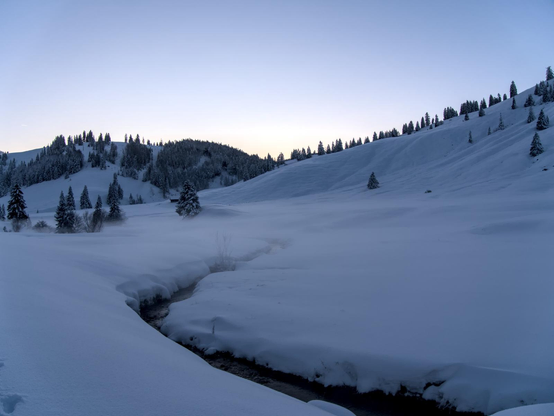 A now coverd valley in the mountains. There is a small stream in the snow. In the background there are some snow covered hills.