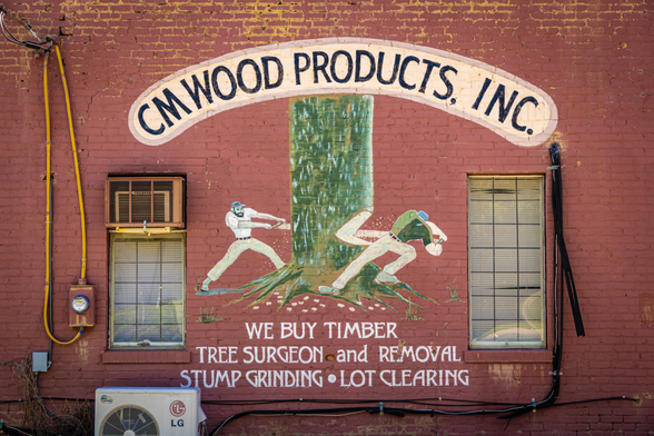 A vintage hand-painted business advertisement on a weathered red brick wall features "CM WOOD PRODUCTS, INC." in an arched cream-colored banner at the top. Below is a folk art mural depicting two lumberjacks using a two-person crosscut saw to fell a large green tree. The text reads "WE BUY TIMBER, TREE SURGEON and REMOVAL, STUMP GRINDING • LOT CLEARING" in white painted letters. The wall has two tall windows with brown frames and venetian blinds. A yellow electrical conduit runs down the left side of the wall with a utility meter, and an LG air conditioning unit is mounted at the bottom left. Various cables and wiring are visible along the bottom of the wall.