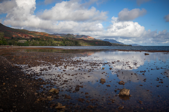 A picturesque coastal landscape featuring a sandy shore with rocks and reflective water, under a partly cloudy sky. Mountains are visible in the background.