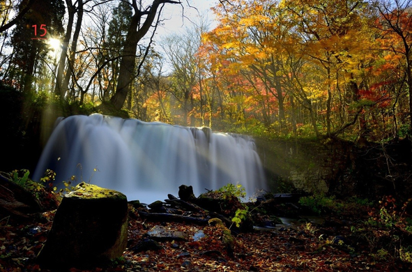 Photo couleur d'une petite cascade pendant l'automne. C'est une exposition longue et l'eau est filée. Devant la cascade des feuilles aux couleurs de l'automne jonchent le sol. Derrière la cascade on voit des arbres aux couleurs de l'automne. Le soleil transperce la couverture arborée qui n'est pas complète à cet endroit.