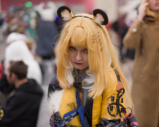 The image features a person in a costume as the central focus. They have long, blonde hair with white tips and bangs, and small brown markings are visible on the hair. Two rounded furry ear accessories (black on top, light brown and white on the inside) are attached to a headband. They wear a yellow jacket with black stripes and leather-like sections. The background is blurred, showing other people in various outfits (e.g., white, black, and brown coats), indicating a busy public setting, likely a convention or event.