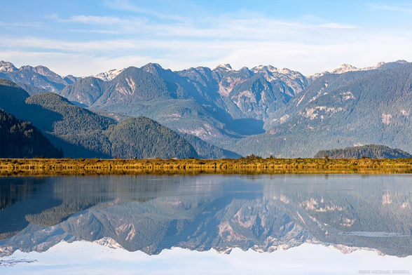 A mountain range with no snow on it in the early fall - is reflected in a foreground marsh with a dike creating a horizontal line between the two across the middle.  The day is sunny, a few clouds in the sky, and the reflection on the marsh is nearly perfect.