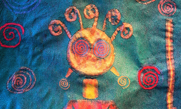 A colorful tapestry made of paper hanging on a wall and illuminated from above by a small, warm spotlight. There is a yellow, orange, and red human figure depicted on the blue-backgrounded tapestry. The human figure is reminiscent of an ancient petroglyph on a rock, and has curly “fronds” coming out its head, plus spiral eyes and earrings, with other red spirals around its head on the blue background.