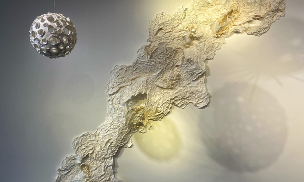 Two more paper sculptures: one attached to the wall, the other hanging from the ceiling. The wall sculpture is made of white paper all scrunched and wrinkled, looking like it’s a river flowing up and down the wall. The ceiling sculpture is a white orb with many holes, looking like a small planet or moon in space. Both are lit so that they cast interesting shadows on the white wall in the background.