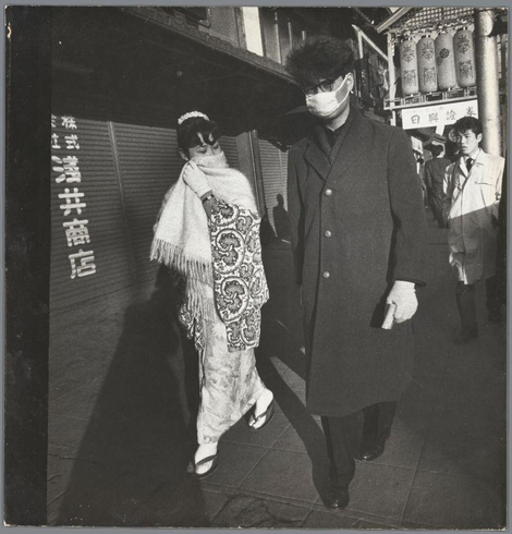 A black and white photograph captures a moment on an urban street. The focus is on two individuals: a man in the foreground, wearing glasses and a dark overcoat with gloves, accompanied by a woman draped in traditional attire featuring intricate patterns of flowers or foliage. She wears sandals typical for such garments. They are walking together; behind them, another person appears partially visible, dressed formally as well. The background is composed mainly of signs, including one written vertically on the left side and other texts that could indicate locations, shops, or public notices in Japanese script, suggesting this setting might be Japan during a time when such modes of dress were prevalent for cultural occasions like weddings.