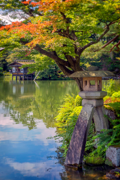 Kenroku-en garden in Kanazawa, Japan. The picture shows a stone lantern at the edge of a lake, surrounded by trees and foliage with a small house on stilts on the shore of the lake in the background.