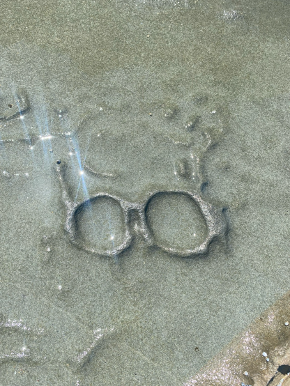 Photograph of a rock formation which looks like a pair of glasses. Sun flare shines on the left side of the picture. The rock is grey-brown with small raised areas above the ‘glasses’