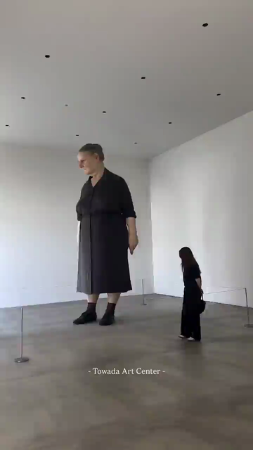 une femme tourne autour de la sculpture hyperréaliste d’une géante monumentale dont le regard la fixe