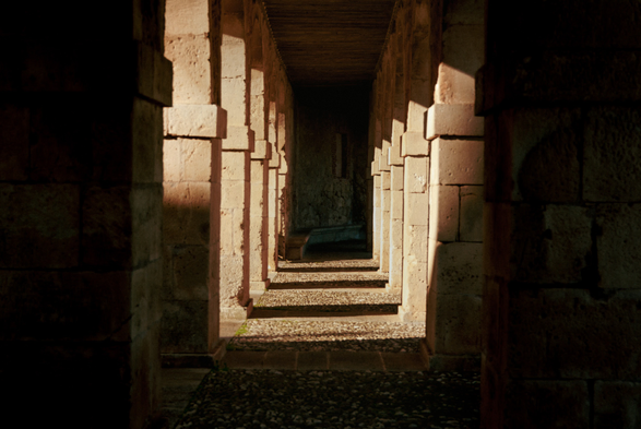 Stone corridor with repeating square columns on both sides, illuminated by warm, angled sunlight that creates alternating bands of light and shadow along the gravel floor, leading toward a darkened depth in the background.