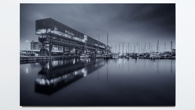 Zwartwit avondopname van Kraanspoor, een bouwwerk waarop grote hijskranen heen en weer konden rijden om schepen te lossen en te laden. Op dit fundament is een kantoorpand gebouwd. Naast dit pand aan het water liggen veel zeilboten. Door de vele lichtjes en het spiegelende wateroppervlak krijgt dit beeld een prachtige dynamiek.