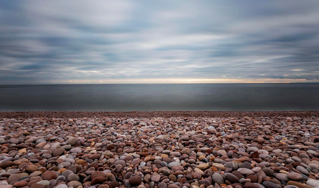 A photo standing on a pebbled beach. The water is flat and dark and blue-gray. The clouds are dark and white and blue. Straight line of pebbled beach leads to straight line of water leads to straight line of clouds. Like a layered cake. Gloomy photo. 