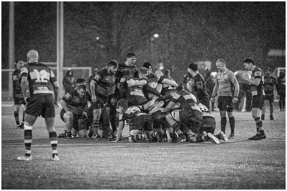 A rugby match is taking place in heavy rain, featuring a group of players clustered in a scrum. The scene is in black and white, with players wearing striped jerseys. A referee and some spectators can be seen in the background.