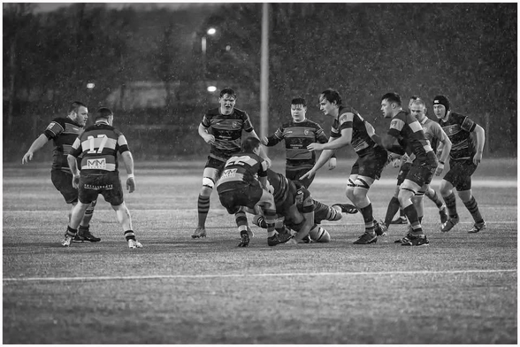 A rugby match in heavy rain, featuring players in striped jerseys engaged in a tackle. The scene is captured in black and white, emphasizing the action and intensity of the game.