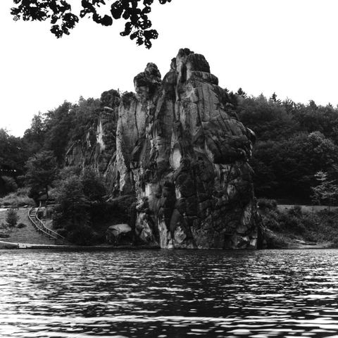 Eine Landschaftsaufnahme in schwarzweiß der Externsteine im Teutoburger Wald. Im Vordergrund ist eine Wasserfläche abgebildet. Neben und hinter den Felsen stehen Bäume.