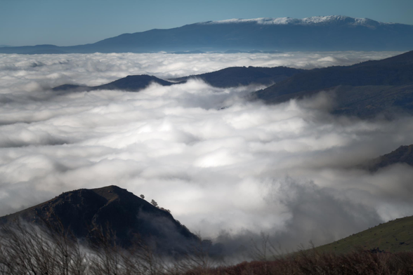 Landscape photo taken from a mountain top above the clouds. There is a dense sea of clouds covering the entire area below. You can't see the bottom of the mountain. The clouds look like a stormy ocean. Another, taller mountain is visible in the distance, as well as a bit of a clear turquoise sky.