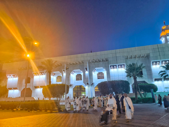 A wide-angle nighttime photograph capturing numerous people dressed in white Ihram robes walking on a paved pathway towards a large, brightly illuminated white building with arched entrances, likely a mosque. The sky above is a gradient of deep blue and purple at dusk. Palm trees and bushes line the walkway. A bright streetlamp on the far left casts a warm lens flare across the frame. A person in a wheelchair is among the pilgrims in the foreground.