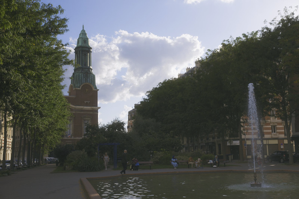 Photo format paysage du centre ville de Bois-Colombe. On y voit au premier plan un morceau de grand bassin probablement carré, avec une fontaine au centre, au second plan, des gens assis sur des bancs et au fond, un bâtiment avec un clocher, probablement la mairie. Des rangées d’arbres forment un cône vers la mairie, le ciel est bleu avec de gros nuage légèrement surexposés