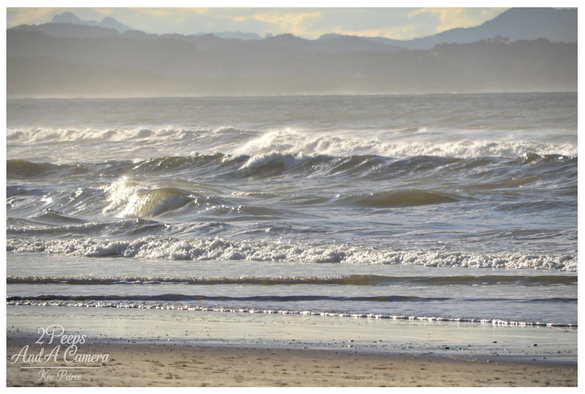 A sweeping, elevated view of a sandy beach on the Northern Rivers coast of New South Wales (NSW), south of Byron Bay. 

Powerful white waves roll in from the deep blue ocean, meeting the golden sand. Coastal scrub and a wooden fence line the foreground, with a distant rock breakwater visible.