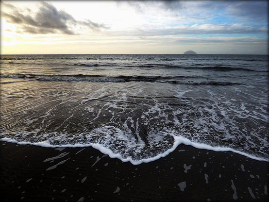 A colour photograph showing the remains of a wave wash ashore on Girvan beach, South Ayrshire. Low afternoon light subdues the colour pallet and provides atmosphere to the image, looking west towards Ailsa Craig in the distant horizon.