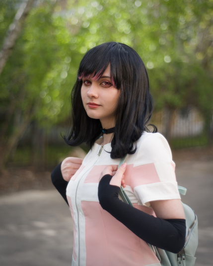The picture shows a person dressed in a costume that appears to be inspired by an anime or manga character. The individual is wearing a long, dark wig that falls just past the shoulders. The outfit includes a white and pink jacket with a zipper and black long-sleeve gloves. Also there is a light-gray backpack behind her back.
The background features a blurred natural setting with greenery and trees, suggesting an outdoor environment. The overall style and details of the costume and accessories indicate a cosplay or character portrayal.