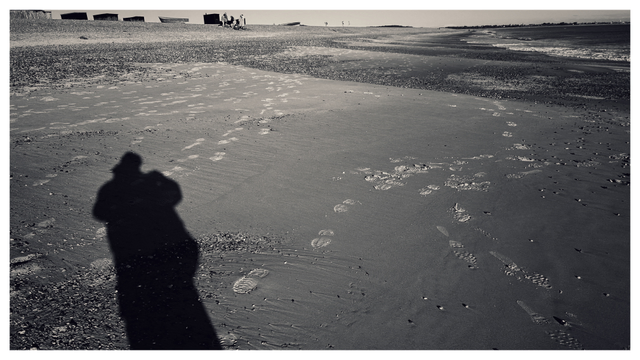 Black and white photo of a long human shadow and footprints on the sand beach leading towards the shoreline with four fishing boats on the left hand side