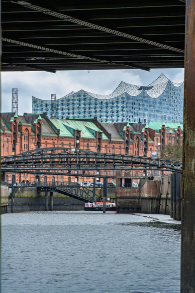 Diese Perspektive bietet einen spannenden Kontrast zwischen Alt und Neu, gesehen unter einer Brückenkonstruktion hindurch.
Historische Speicherstadt: Im mittleren Bereich dominieren die klassischen Rotklinker-Speicher mit ihren grünen Kupferdächern und typischen Giebeln.
Die Elbphilharmonie: Über den historischen Speichern thront die moderne Silhouette der Elbphilharmonie, was den architektonischen Wandel der Stadt verdeutlicht.
Wasserwege & Brücken: Im Vordergrund sieht man eine der vielen Hamburger Stahlbrücken, die über einen Fleet (Kanal) führen, auf dem eine kleine Barkasse fährt.